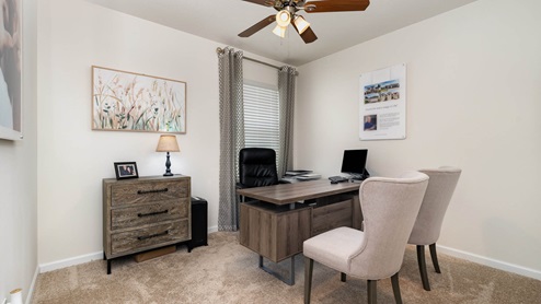 A cozy home office with a wooden desk, black swivel chair, two plush light gray chairs, lamp, and decorative artwork on the walls.