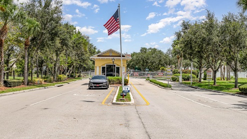 Entrance to a gated community with American flag, palm trees, and a parked car, under a bright blue sky.