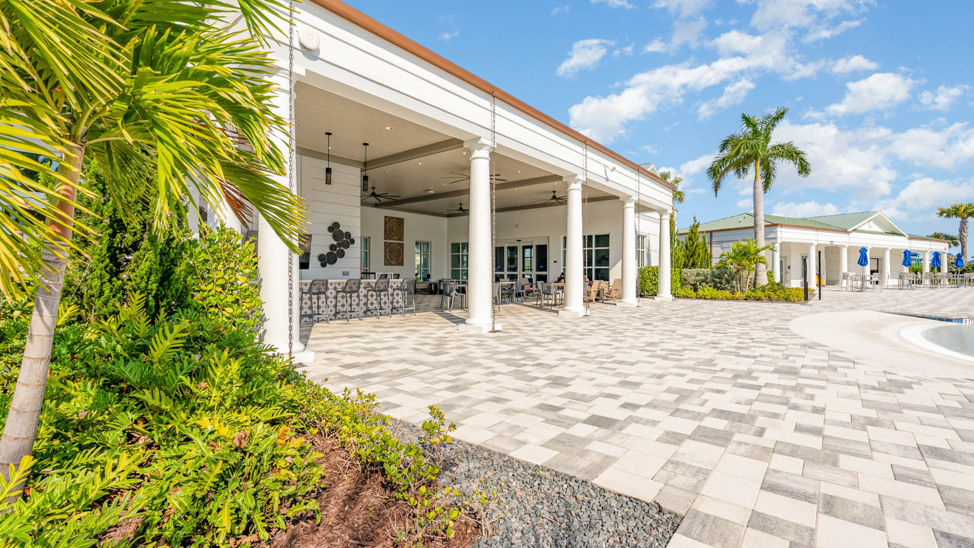 Spacious patio with white columns and modern seating, bordered by lush green plants. A pool is visible, set against a bright, sunny sky.