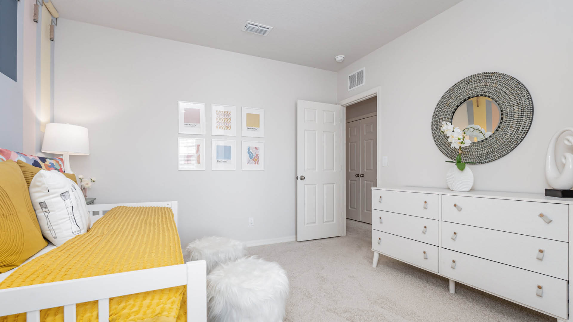 A cozy bedroom with a white bed featuring a yellow blanket and pillows. The room includes a white dresser with a round mirror above, soft stools, and wall art, creating a calm, modern vibe.