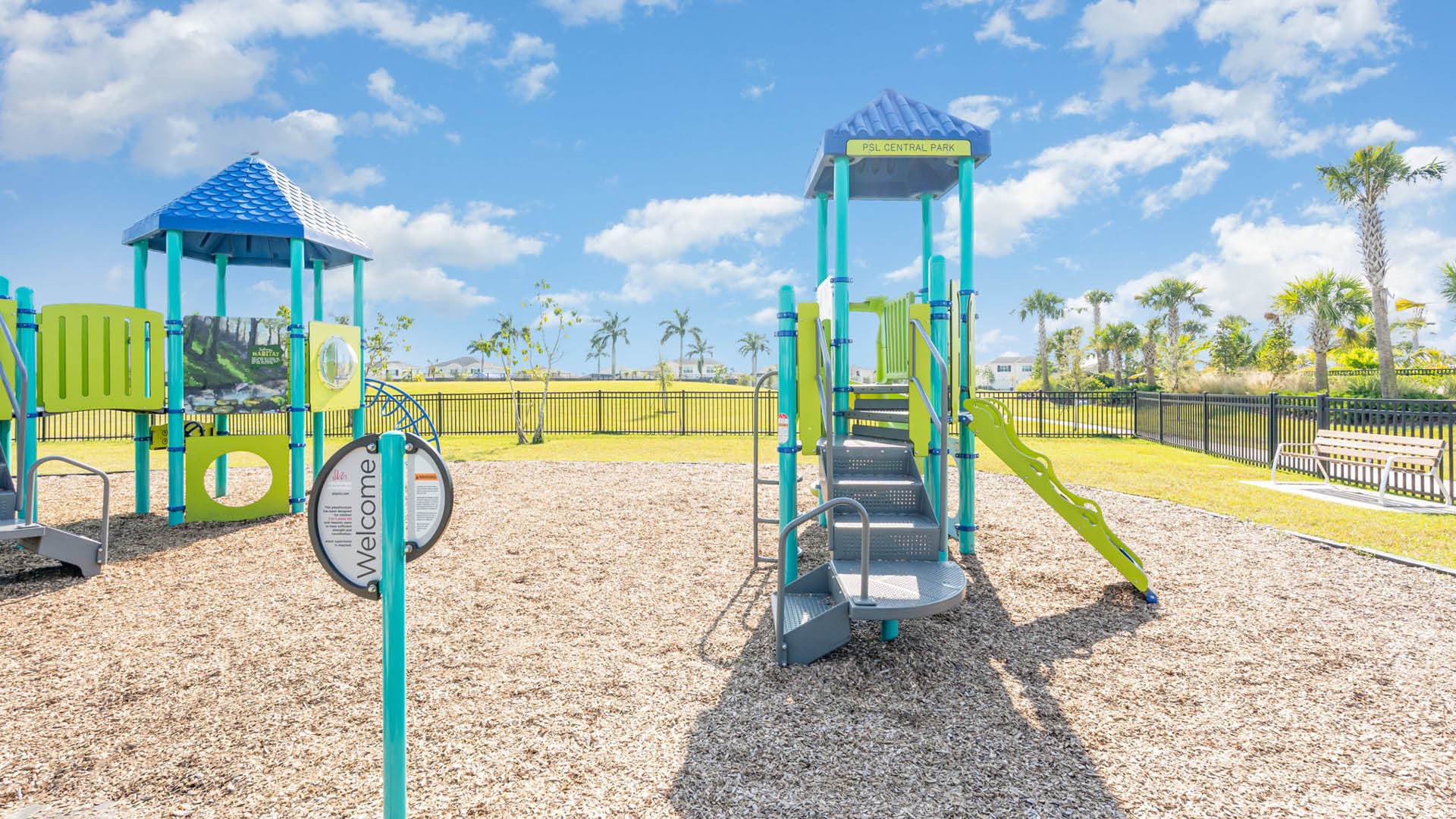 Sunny playground with blue and green equipment, including slides and climbing structures on wood chips. Palm trees and a bench are in the background.