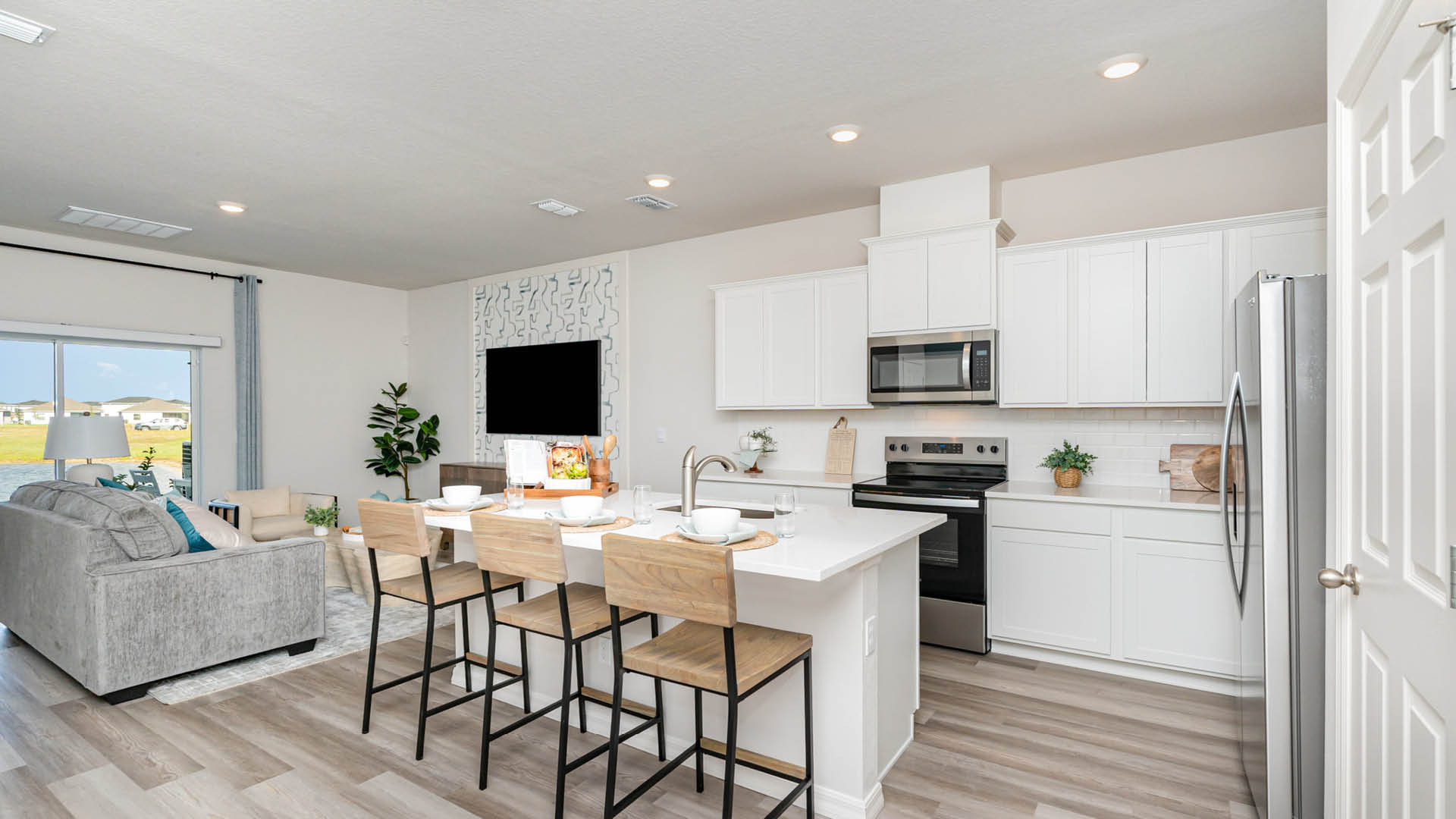 A modern kitchen and living area with a white color scheme, featuring a kitchen island, stainless appliances, and large windows.