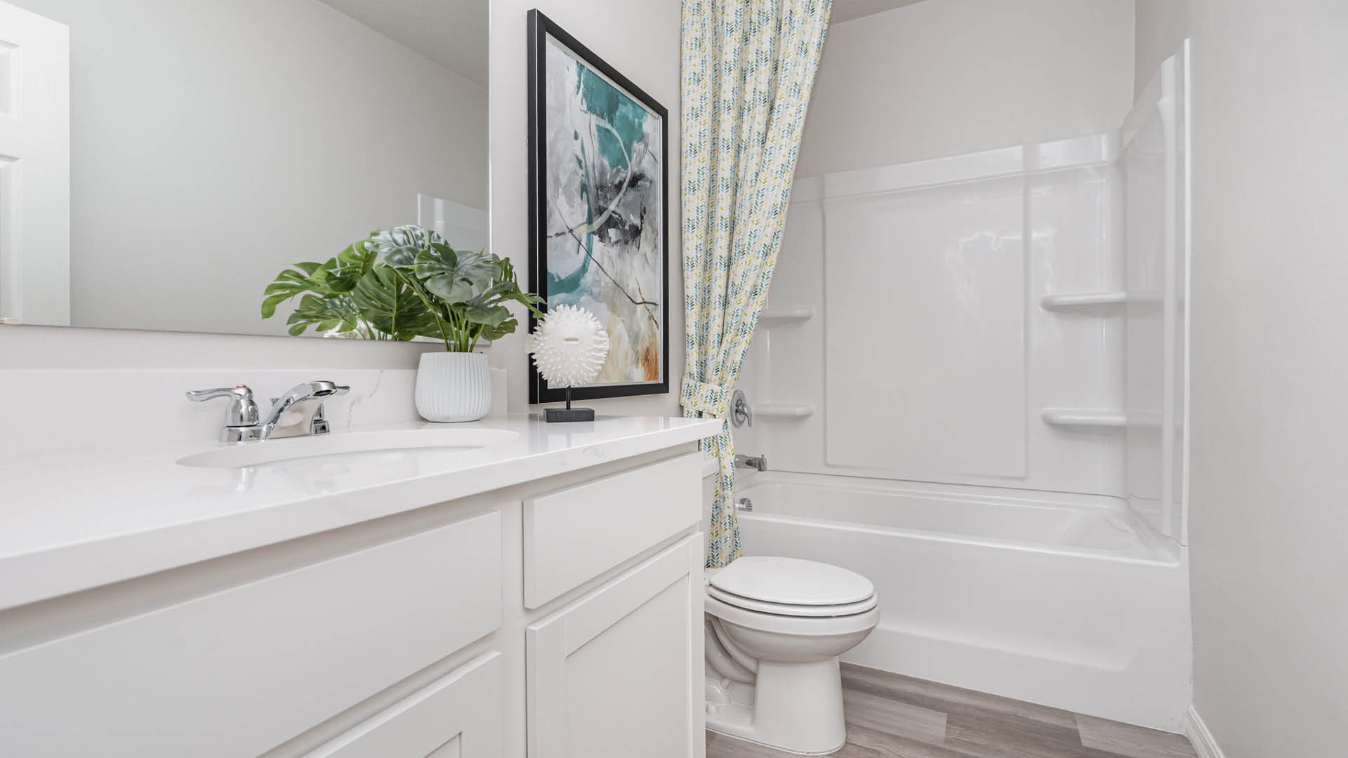 White bathroom with a modern, clean look. Features a vanity with a plant, abstract art, floral shower curtain, and a tub, exuding a serene ambiance.