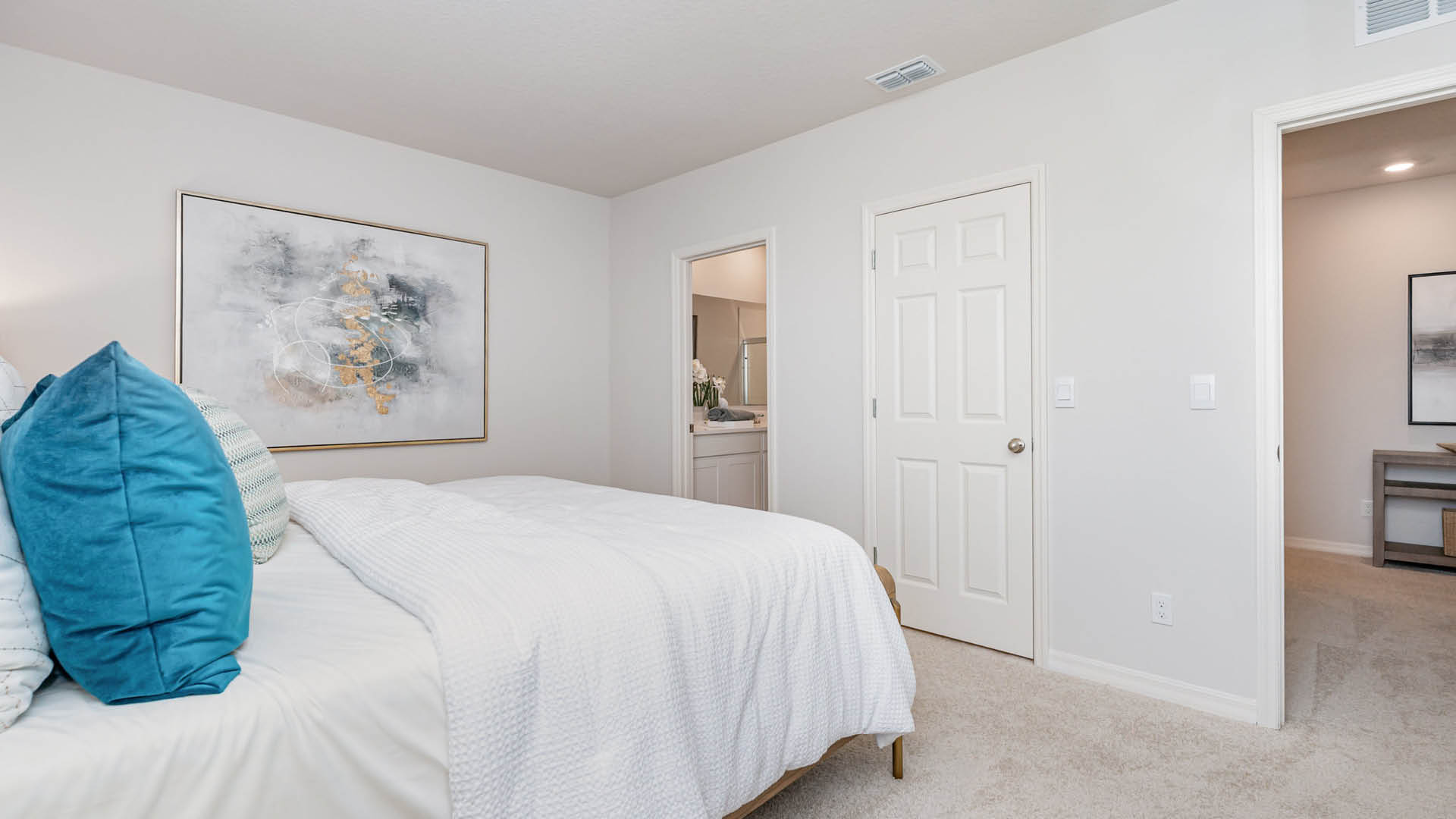 Bright bedroom featuring a white bedspread, colorful cushions, an abstract wall art, and an open bathroom door in a cozy setting.