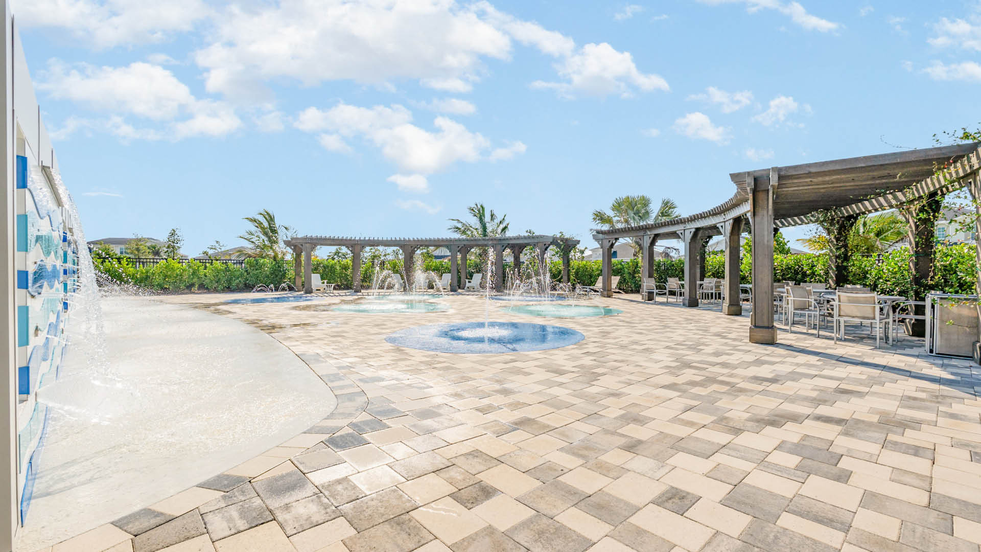 Sunny outdoor splash pad with fountains, surrounded by a spacious tiled area. Wooden pergolas with seating provide shaded spots, under a blue sky.