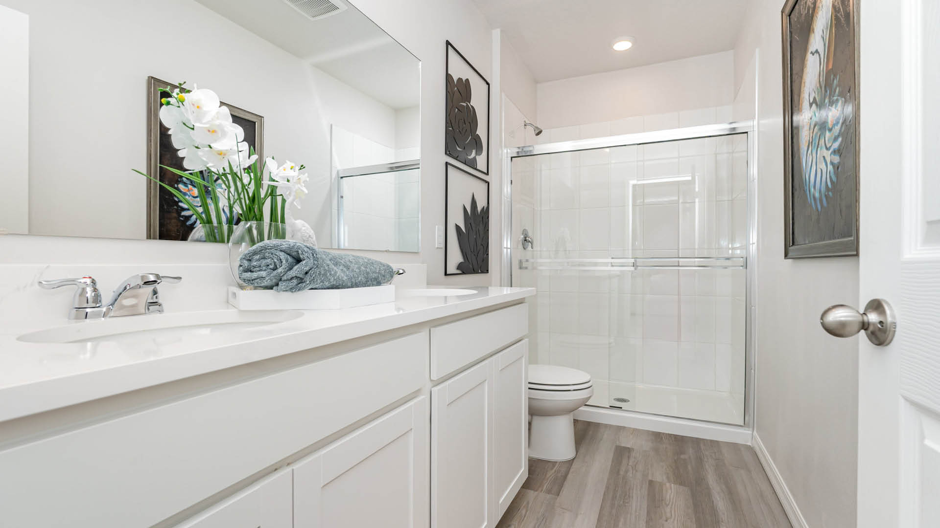 Modern bathroom with white cabinets, double sink, glass shower, toilet, vinyl pank flooring, wall art, and a vase with white flowers on the counter.