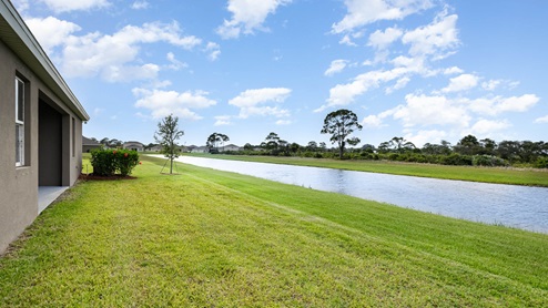 Lush green lawn beside a tranquil waterway, with bright flowers and trees, under a clear blue sky. Residential homes in the background.