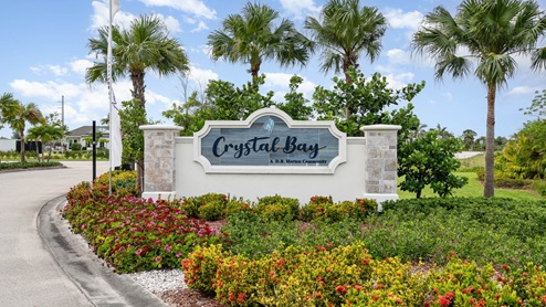 Welcome sign for Crystal Bay, a D.R. Horton community, surrounded by tropical plants and colorful flowers against a clear blue sky.