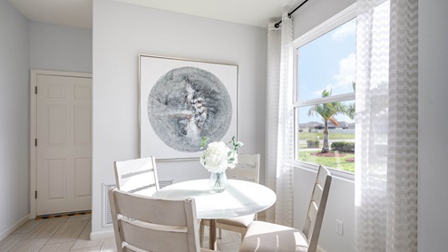 Bright dining area featuring a round table, vase of white flowers, and a large circular wall art, with sunlight streaming through a window.