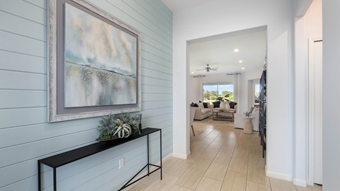 Bright entryway with light blue shiplap walls, a framed painting, and a modern table, leading to a cozy living room with natural light.