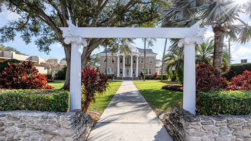 Elegant two-story stone house with white columns, surrounded by vibrant greenery and flowers, seen through a decorative archway.