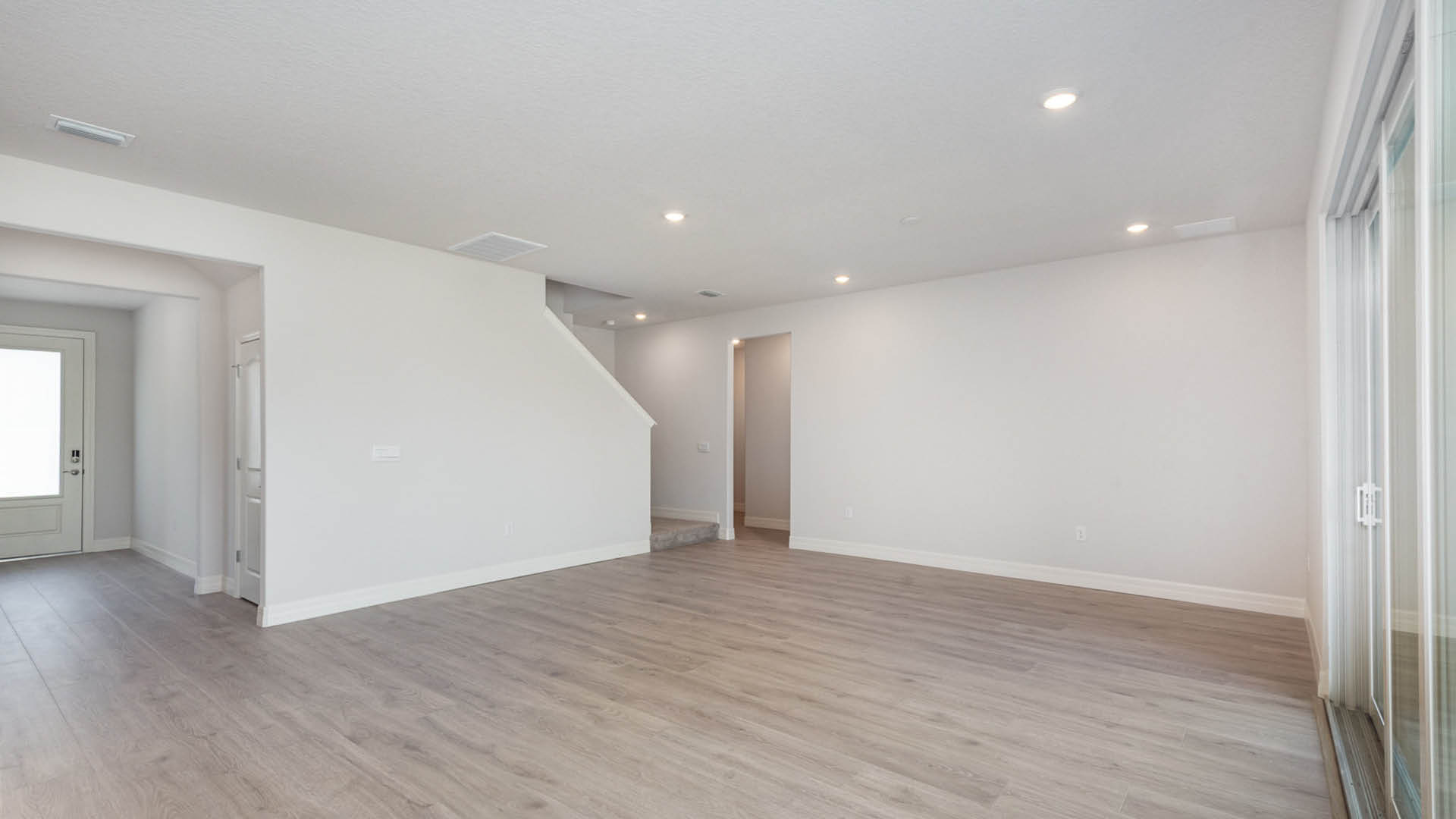 Spacious, empty living room with light gray walls, vinyl plank flooring, and natural light streaming through sliding glass doors.