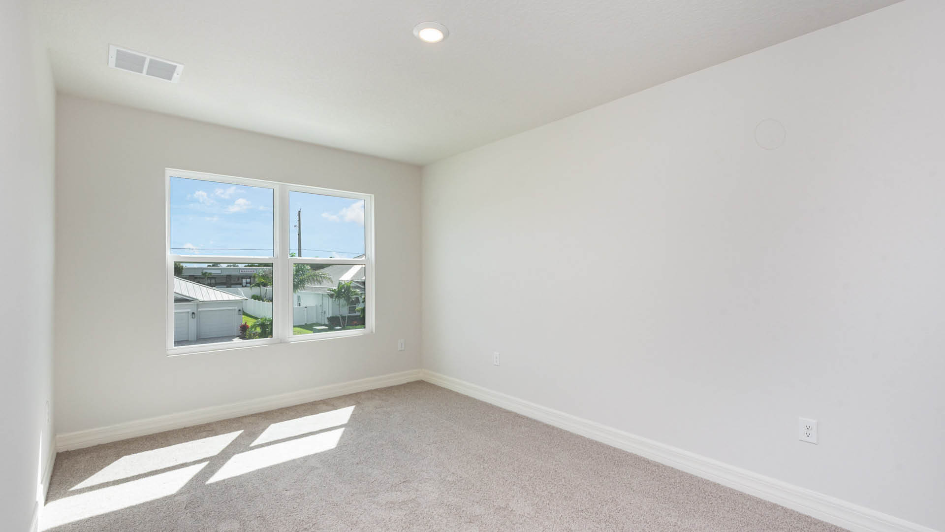 Bright, empty room with beige carpet, white walls, and a large window showing a blue sky and neighboring houses outside.