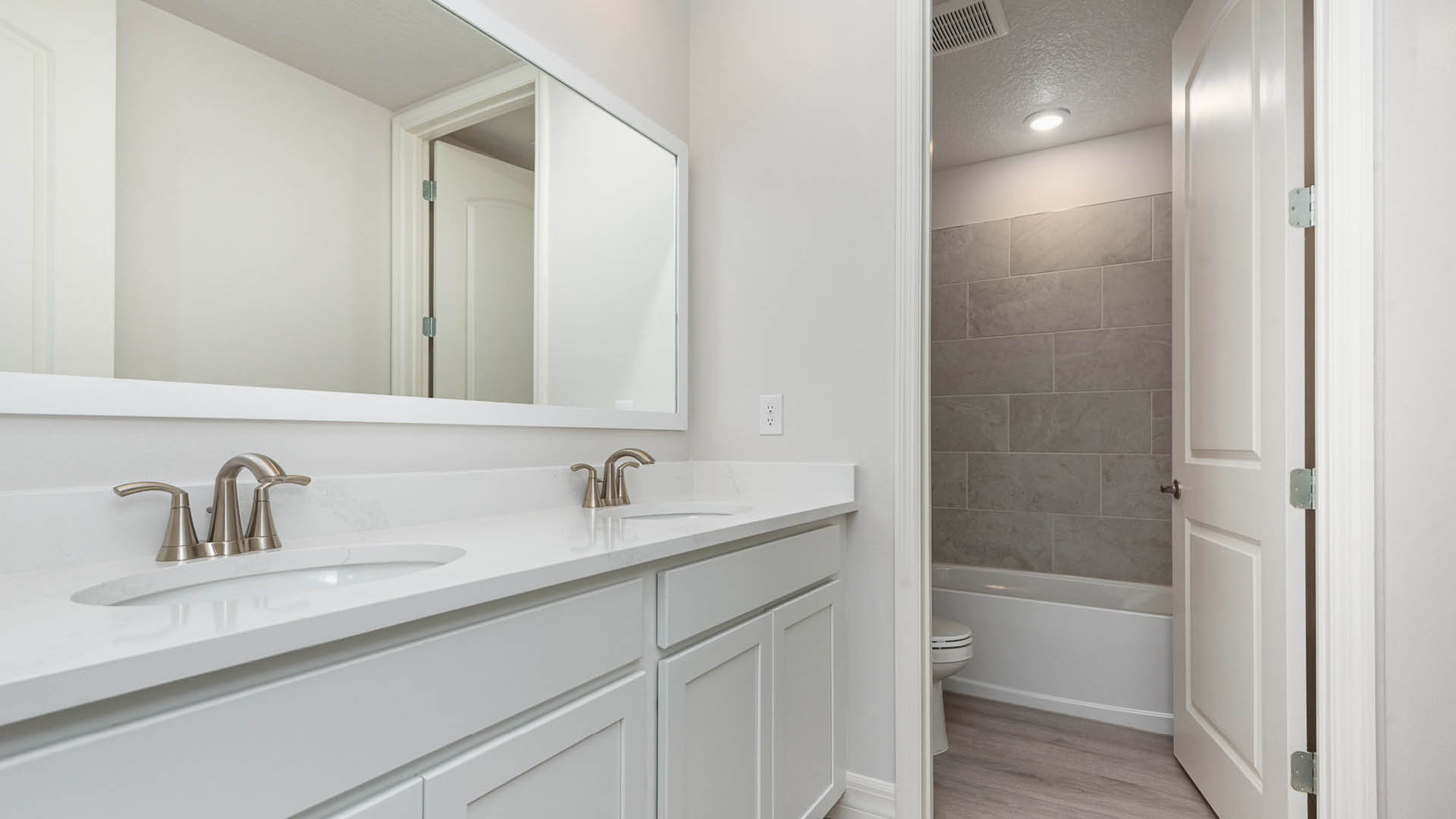 Modern bathroom with dual sinks, a large mirror, and an adjoining room featuring a shower and toilet. Light, neutral colors throughout.