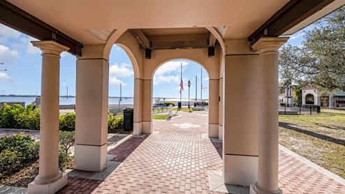View through a stone archway, offering a glimpse of a waterfront park with benches, flags, and a clear blue sky.