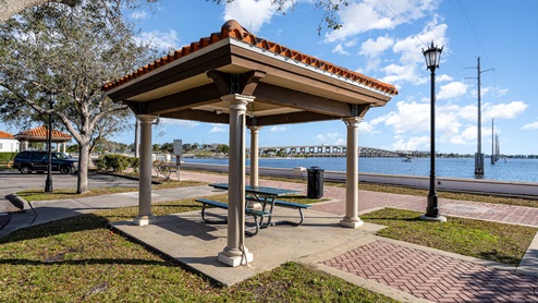 A gazebo with a terracotta roof and picnic table overlooks a waterfront, with a bridge.
