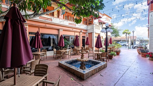 Outdoor dining area featuring tables with maroon umbrellas, a decorative fountain, and a vibrant, sunny atmosphere.