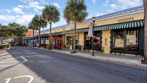 Colorful storefronts with striped awnings line a quaint street, flanked by palm trees and under a bright blue sky.