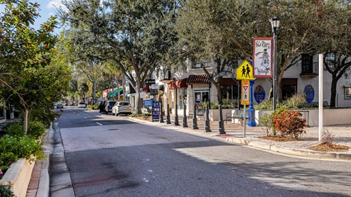 A picturesque street lined with trees and shops, featuring parked cars and vibrant signage under a clear blue sky.