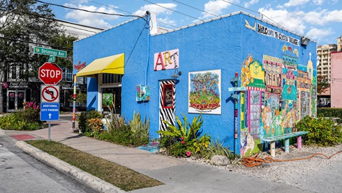 A vibrant blue building with colorful murals and signs, located at Delannoy Ave, featuring an art co-op and lush landscaping nearby.