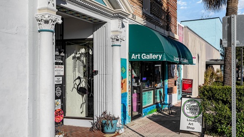 Exterior view of a vibrant local art gallery, featuring colorful murals, potted plants, and a sign for psychic consultations nearby.