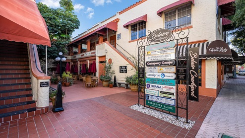 A vibrant courtyard with shops and dining, featuring colorful signage and outdoor seating under red umbrellas in a sunny setting.