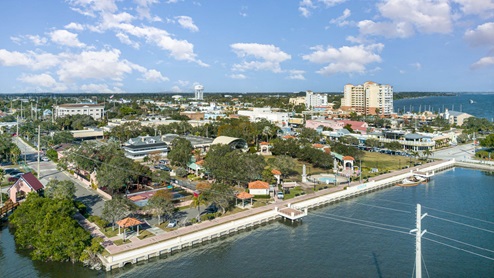 Aerial view of a coastal town with waterfront parks, colorful buildings, and a marina under a clear blue sky.