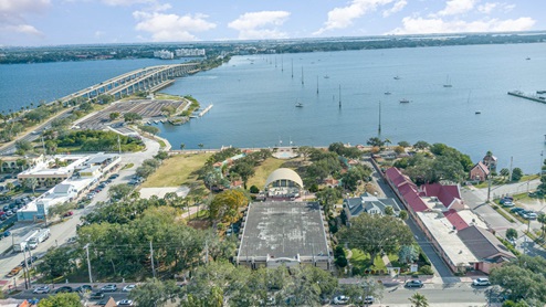 Aerial view of a waterfront park with a bandstand, nearby marina, and retail shops, framed by a bridge and the waterway beyond.