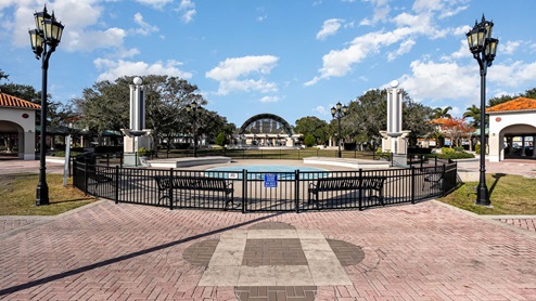 A serene park scene featuring a fenced fountain, benches, and decorative lamp posts.