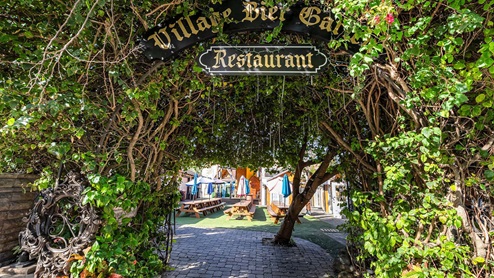 An entrance archway covered in greenery leads to a cozy outdoor dining area of "Village Bier Garten" restaurant.