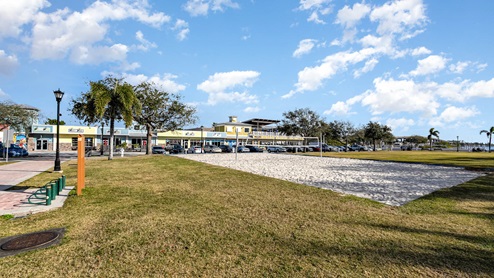 A sunny park featuring a sandy volleyball court, colorful shops, and cars parked along a grassy area.