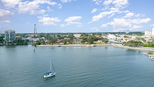 A scenic view of a waterfront area with a sailboat, shops, and lush trees under a partly cloudy sky in a coastal town.