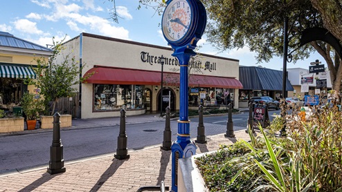 A blue clock stands in front of the Threadneedle Shopping Mall, showcasing vibrant storefronts and clear skies in a quaint town setting.