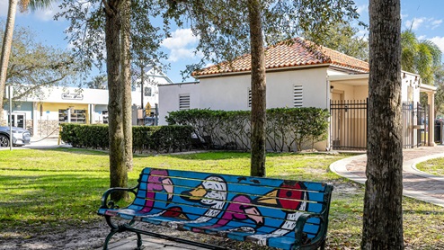 A colorful, painted bench featuring whimsical birds in a park setting, with a building and greenery in the background under a blue sky.