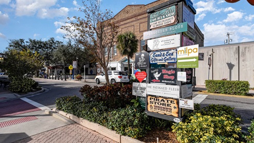 A street signpost displaying various local shops and restaurants in a sunny, quaint downtown area with blue skies and greenery.