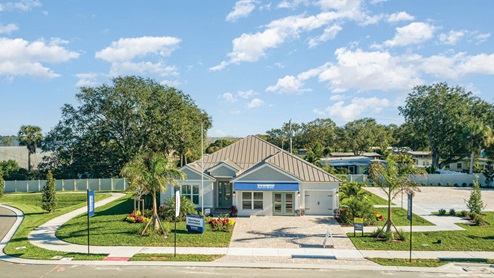 A modern, single-story model home with a metal roof, large windows, and a landscaped front yard sits on a corner lot with signage indicating it is a sales center for new homes.