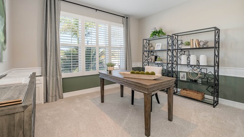 A room decorated as an office with neutral walls on top and dark green at bottom. Light carpet and large window.