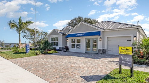 Single-story model home with a metal roof, large windows, and paver driveway with signage indicating it is a sales center for new homes