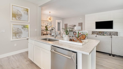 A modern open-concept kitchen features a quartz island with a stainless steel dishwasher, stylish décor, and a view into the dining and living areas with elegant lighting and neutral tones.
