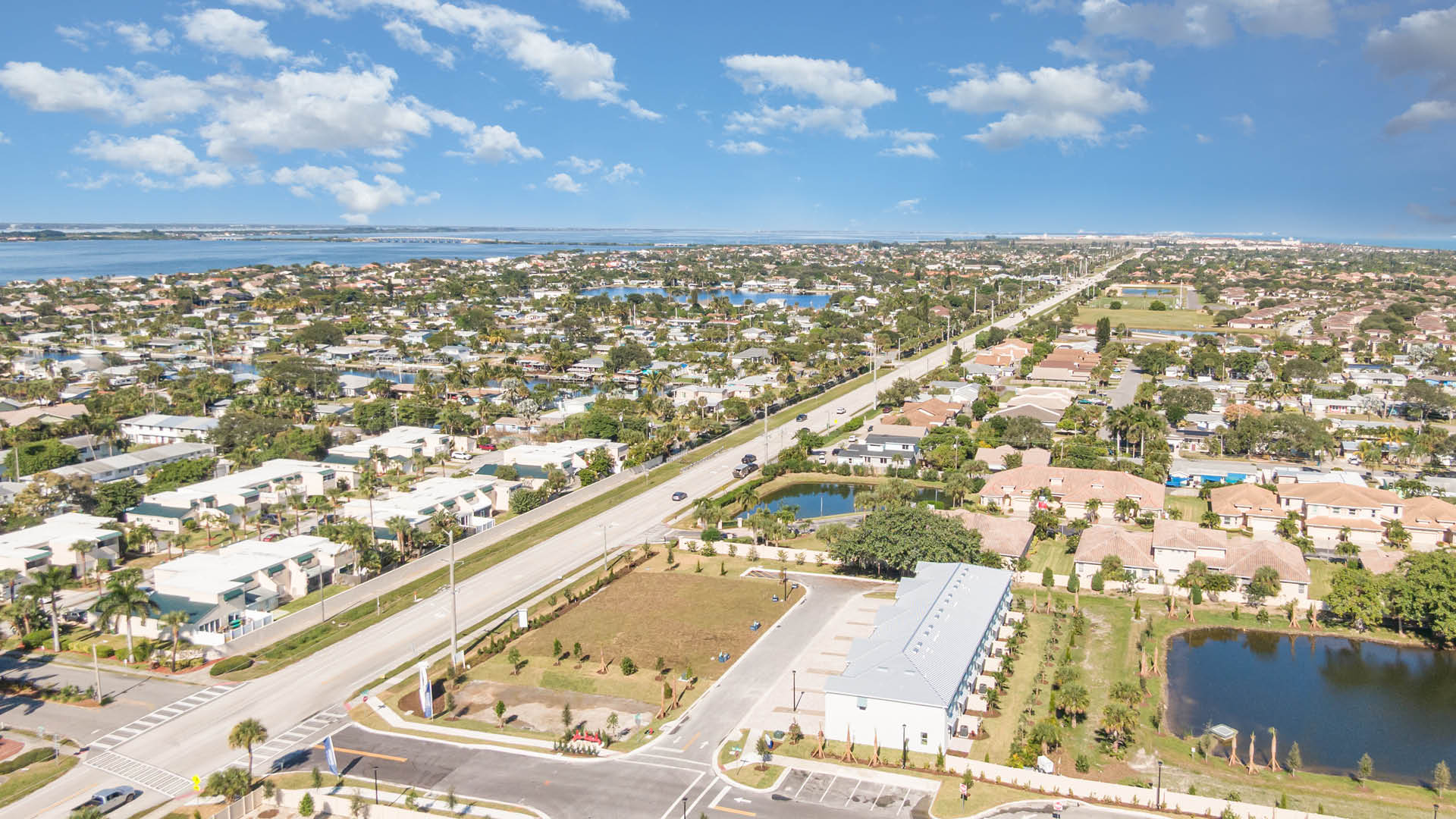 Aerial view of a Tortuga Cay featuring townhomes, a pond, commercial areas, and the ocean in the distance under a blue sky.