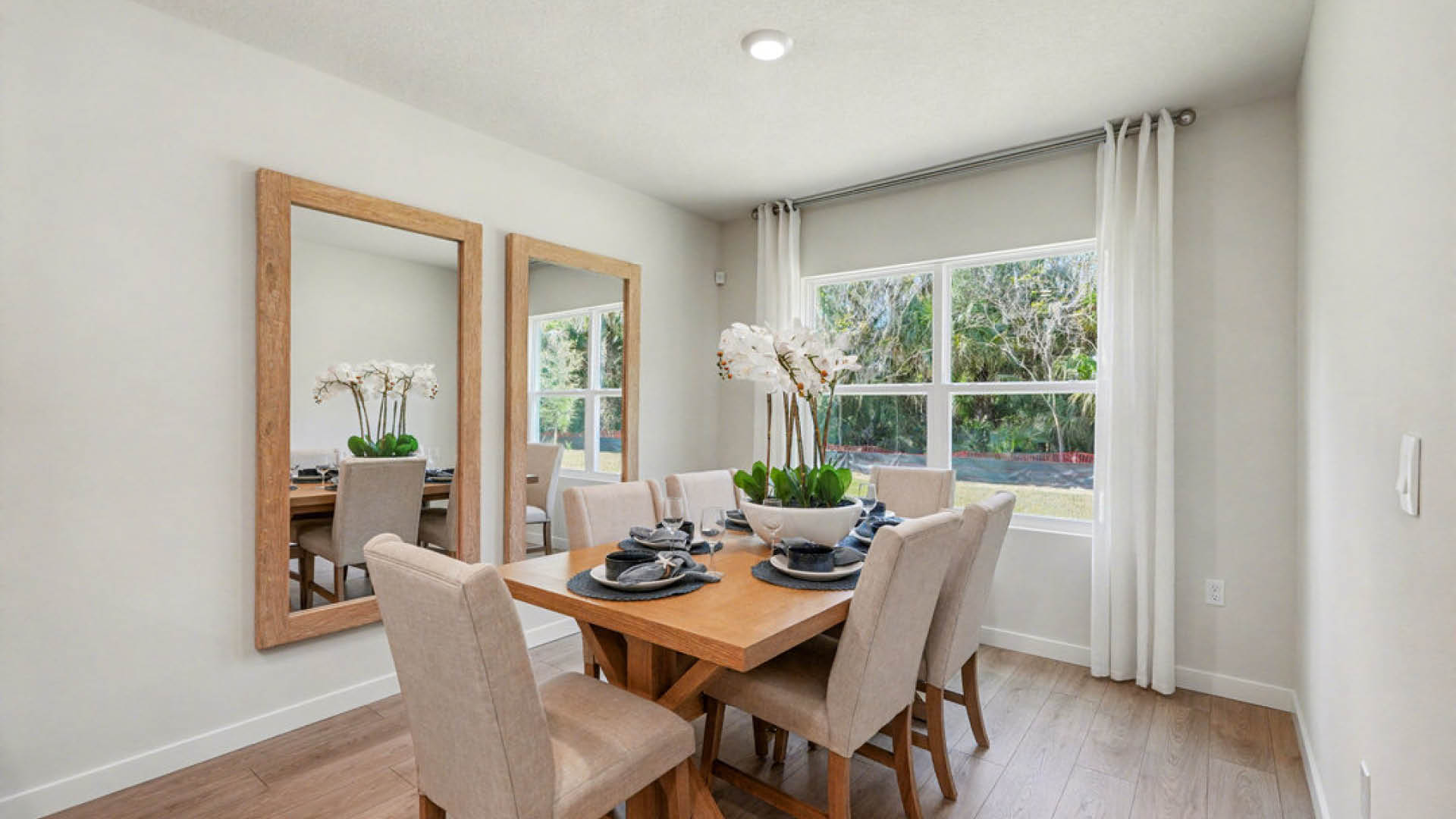 Bright dining area featuring a wooden table, beige chairs, and large mirrors, accented with a vase of orchids and natural light.