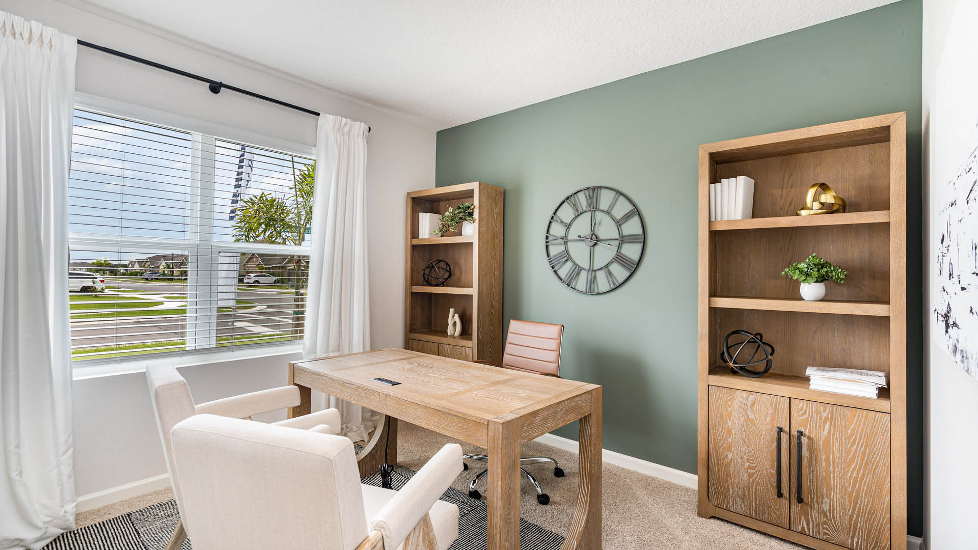 A modern home office with a green accent wall features a wooden desk, two beige chairs, and bookcases. Large window with white curtains showing a suburban view.