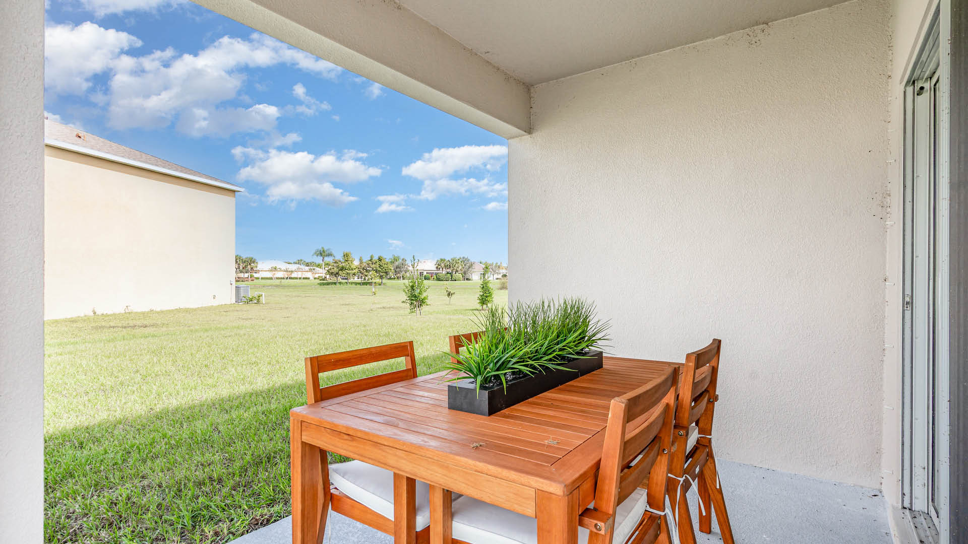 Covered patio with a wooden table and chairs, adorned with a rectangular plant centerpiece. Overlooks a sunlit grassy yard under a bright blue sky.