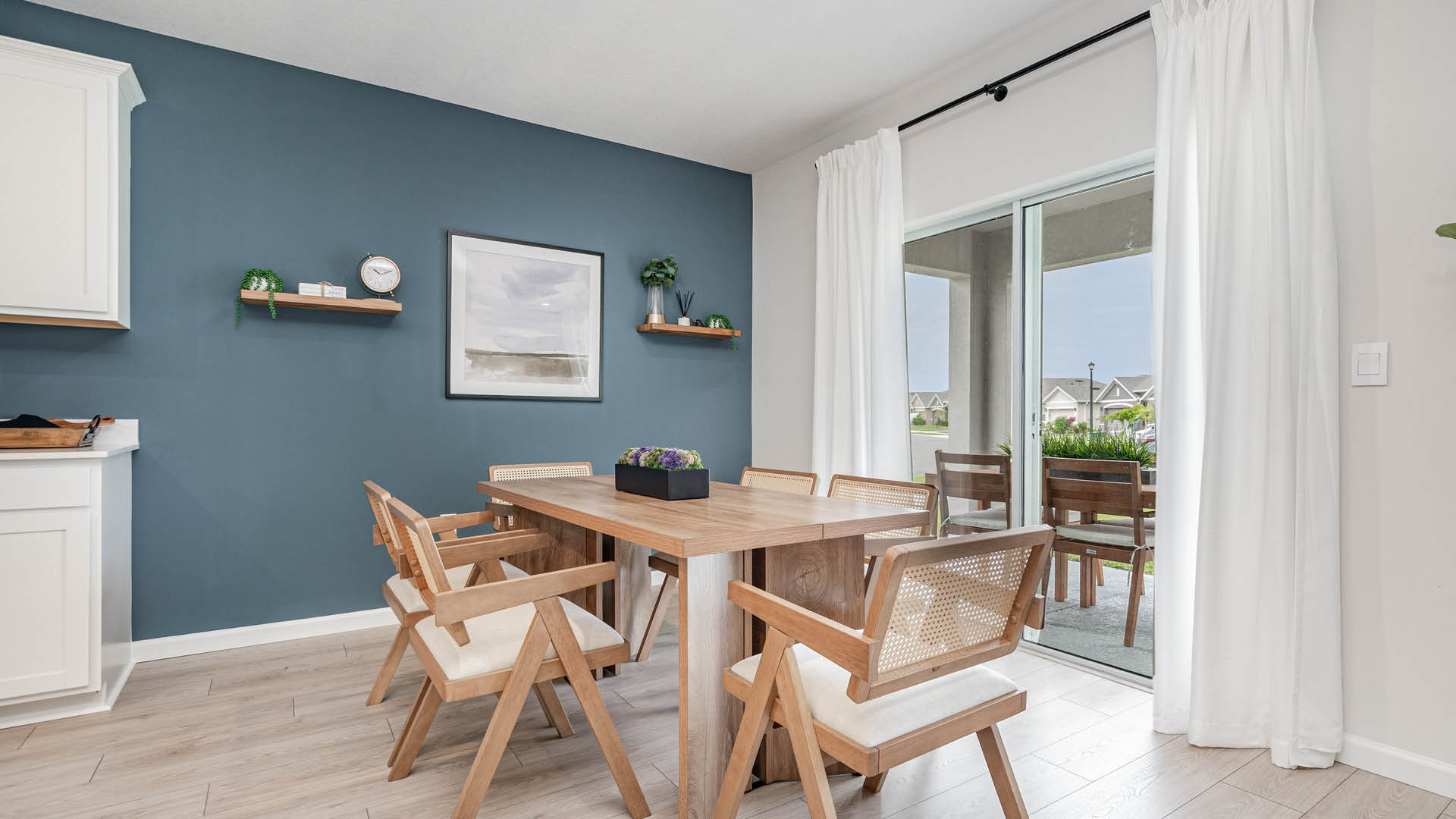 A modern dining area featuring a wooden table, eight chairs, and a blue accent wall with decor and natural light from large sliding glass door.