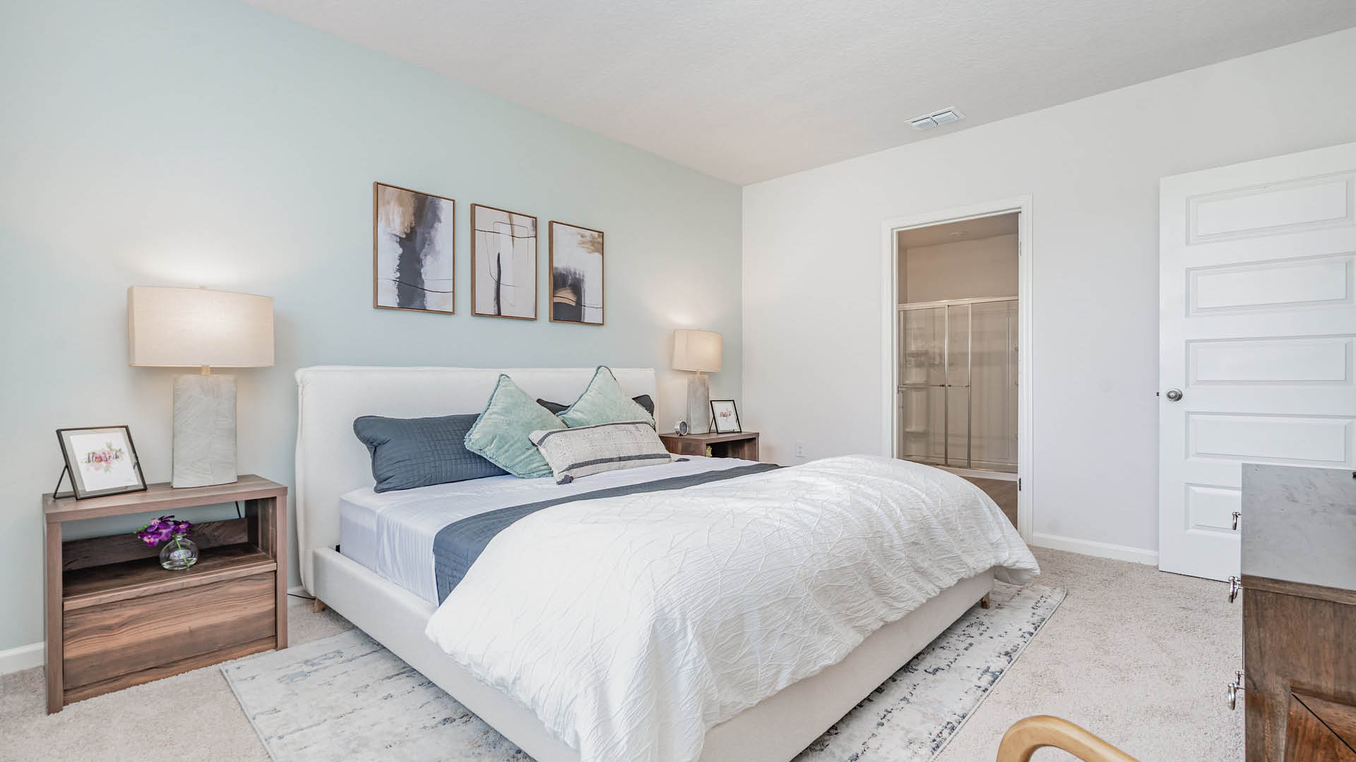 Modern bedroom with a large white bed adorned with gray and green pillows, flanked by wooden nightstands and lamps. Soft pastel walls and abstract art create a tranquil ambiance.