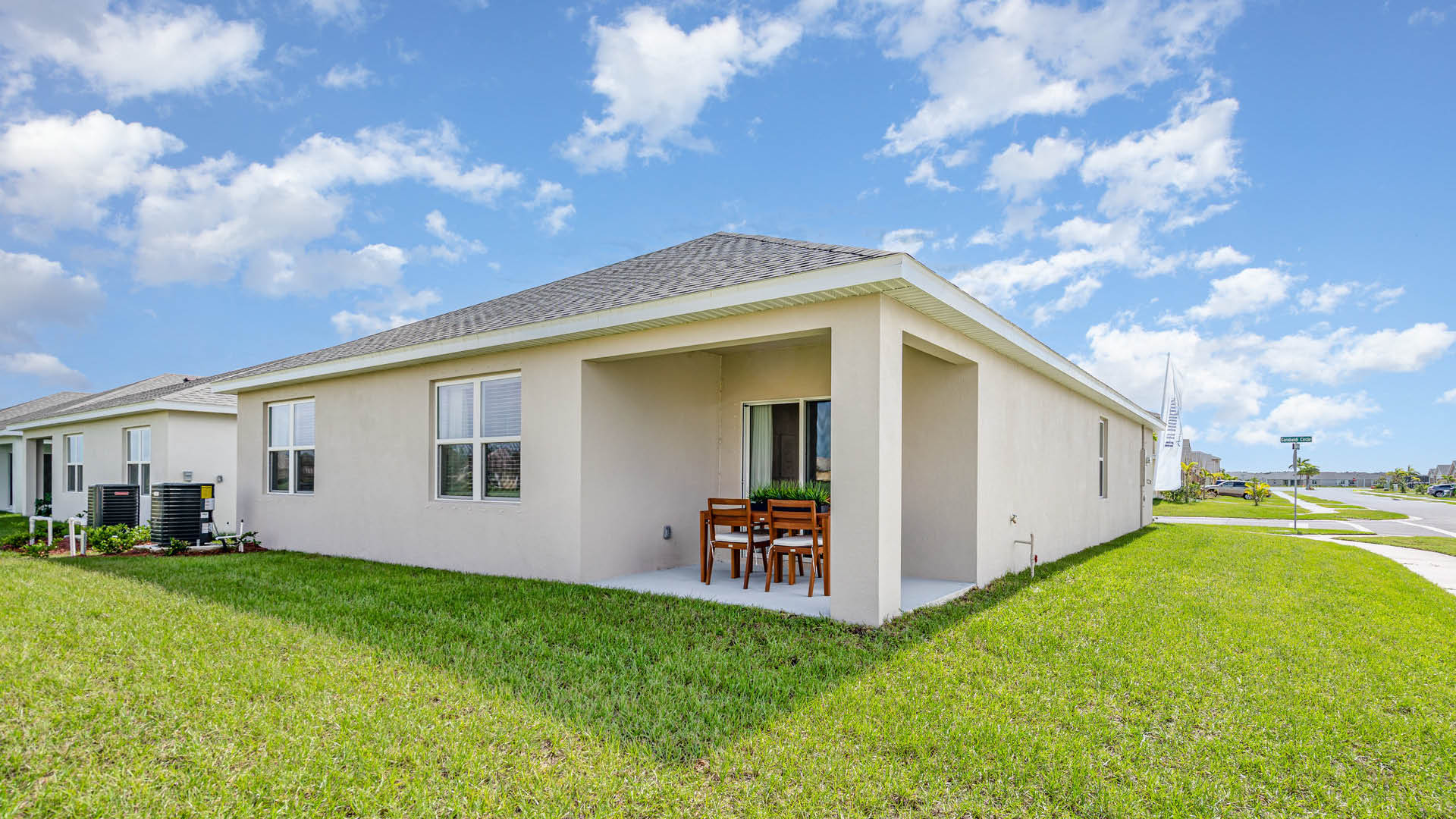 Single-story beige house with a small patio set, surrounded by a green lawn. Bright blue sky with scattered clouds, creating a serene atmosphere.