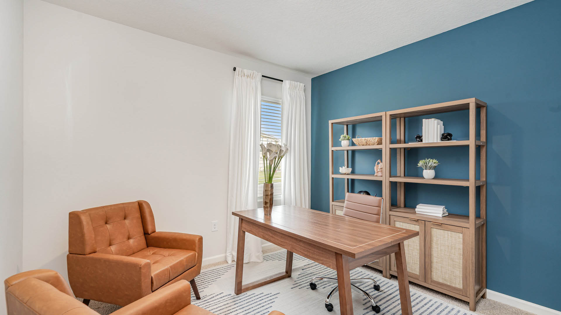 Modern home office with leather chairs, wooden desk, and blue accent wall. Shelves display books and plants, creating a cozy, minimalist atmosphere.