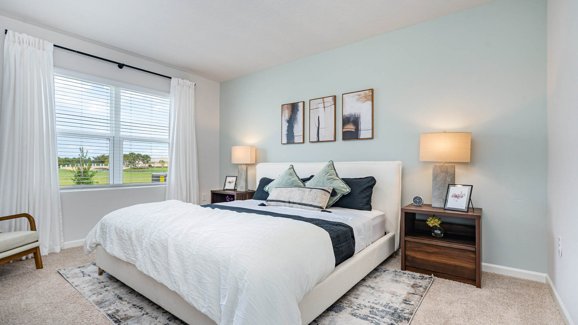 A serene bedroom with a large bed featuring white and navy bedding, flanked by wooden nightstands with lamps. Light blue walls and a window with curtains let in natural light, creating a calm atmosphere.