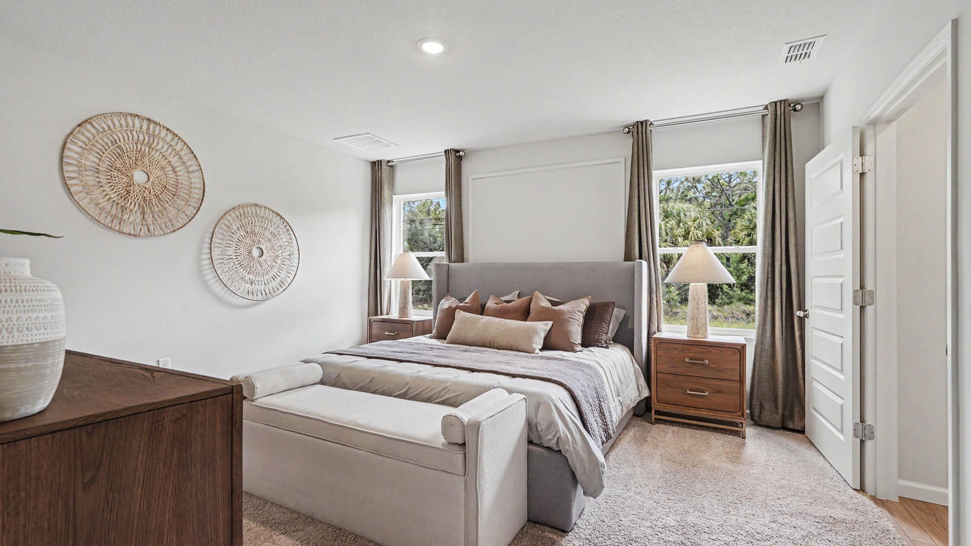 A large bedroom with beige carpet, white walls, and two windows, allowing natural light to illuminate the space. An open door leading to an ensuite bathroom is visible.