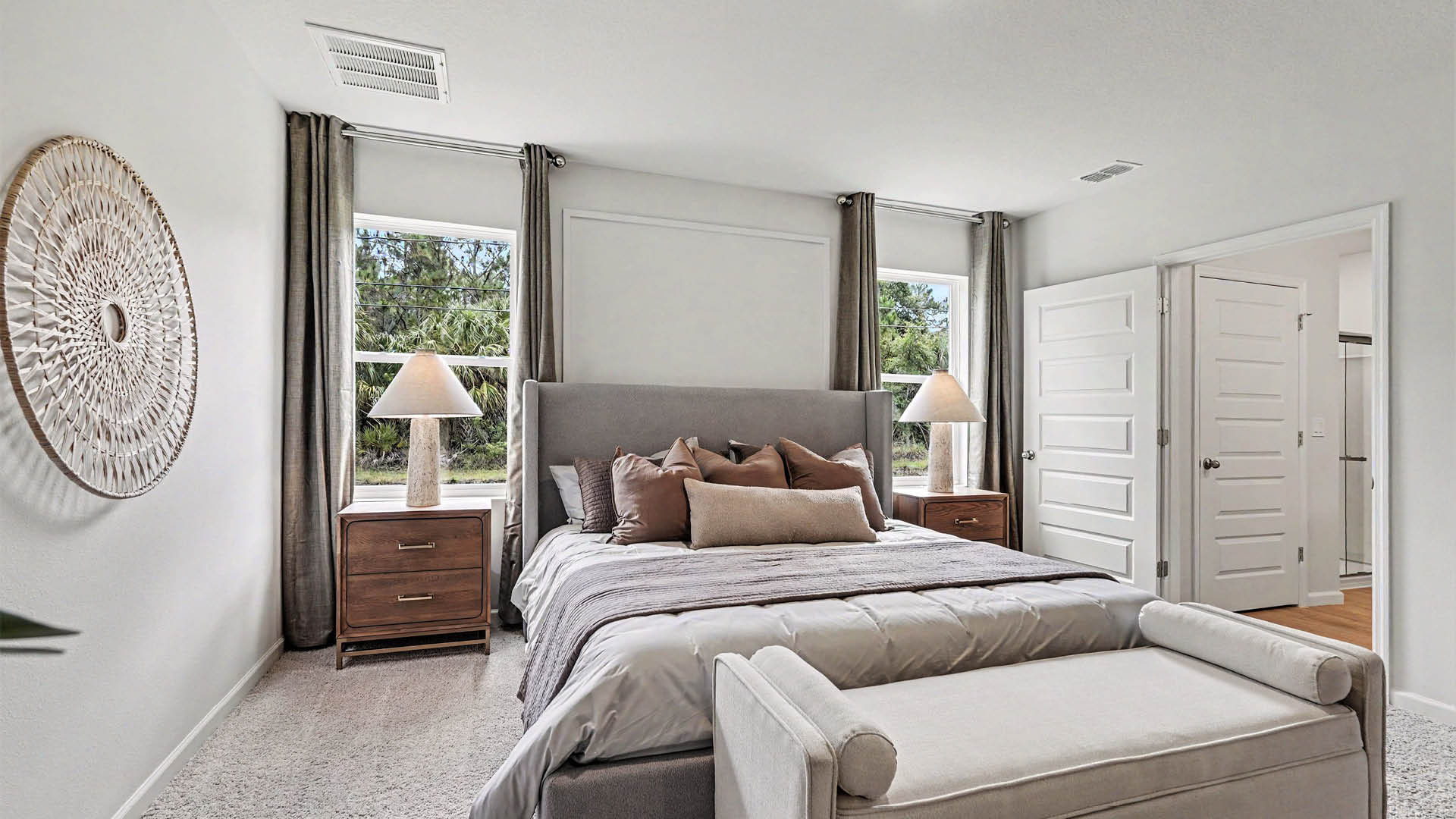 A large bedroom with beige carpet, white walls, and two windows, allowing natural light to illuminate the space. An open door leading to an ensuite bathroom is visible.